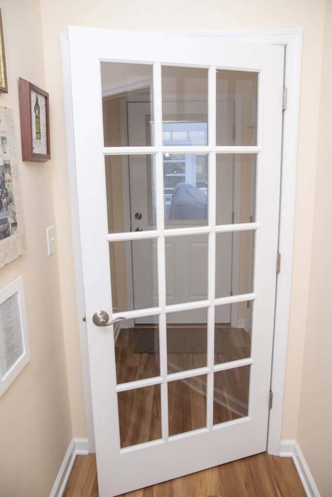 A white French door with glass panes and a silver handle opens into a small entryway with light-colored walls and wood flooring; another white door is visible in the background.