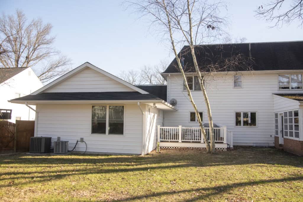 A white two-story house with a small wooden deck, bare trees, and an open grassy backyard. The house has black roof shingles, multiple windows, and two HVAC units on the left side.