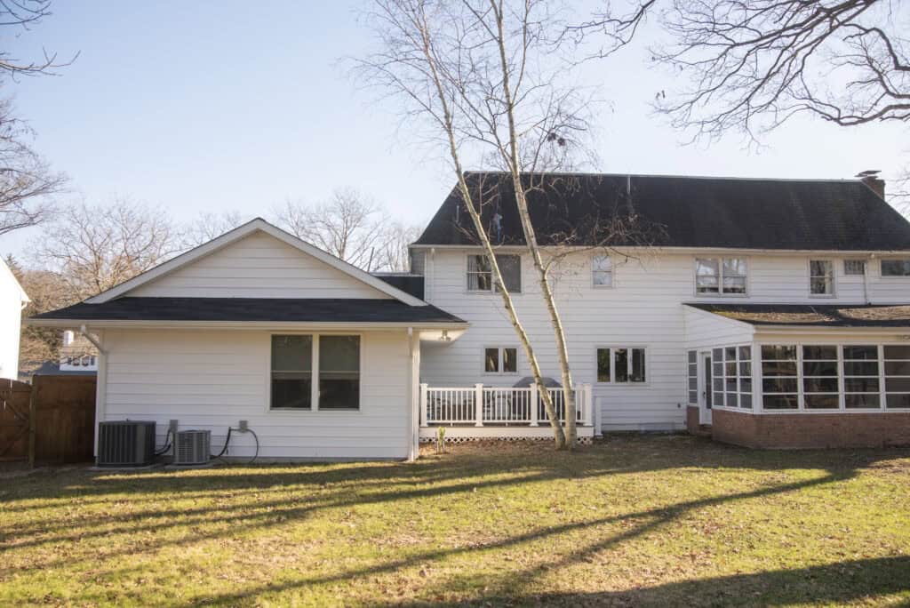 A white two-story house with a small porch, an attached sunroom, two air conditioning units, and leafless trees in a grassy backyard on a sunny day.
