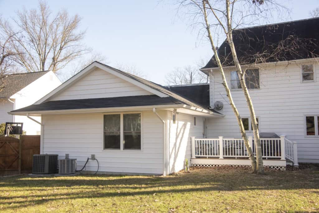 A white house with a small wooden deck, two air conditioning units, leafless trees, and a grassy backyard on a sunny day.