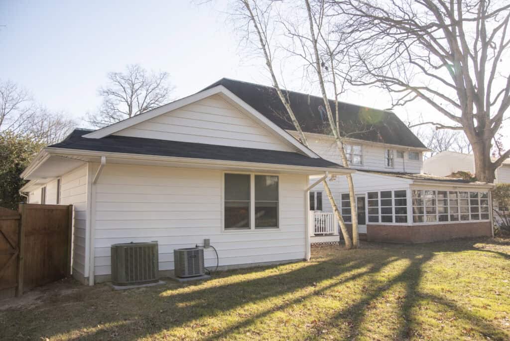 A white house with horizontal siding, black roof shingles, a fenced yard, two outdoor HVAC units, and leafless trees casting long shadows on the lawn under a clear sky.