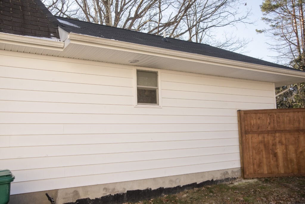 A white house with horizontal siding, a small window near the center, a wooden gate on the right, a green garbage bin on the left, and bare trees visible above the roofline.