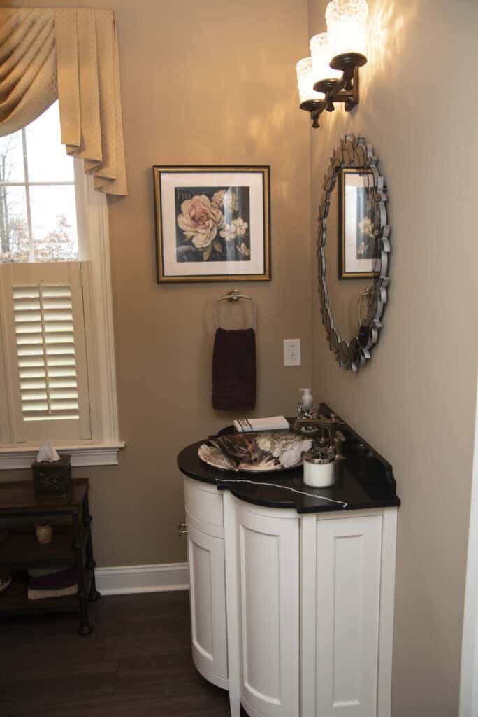 A small bathroom with a white vanity, black countertop, decorative mirror, wall sconce, framed floral artwork, towel ring with a dark towel, window with shutters, and a shelf with tissues and decor.