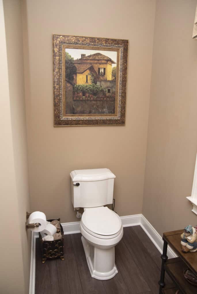 A small bathroom with beige walls, featuring a white toilet beneath a framed painting of a house, a basket holding extra toilet paper, and a window with natural light on the right.