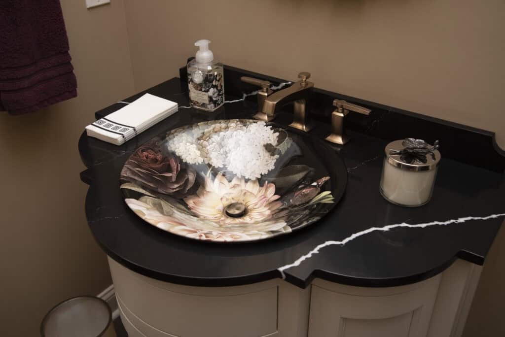 A bathroom vanity with a black marble countertop, a floral-patterned sink, bronze faucet, soap dispenser, folded napkins, and a candle jar, against a warm beige wall.