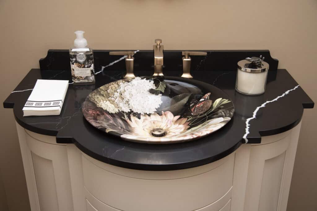 A bathroom sink with gold fixtures, a decorative floral-patterned basin, black countertop with white veins, a soap dispenser, folded towels, and a candle jar arranged neatly.