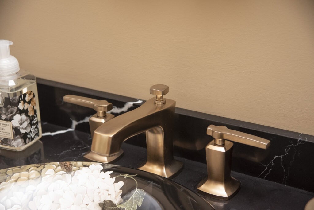 A bathroom sink with a bronze faucet and handles on a black marble countertop, a decorative bowl with white flowers, and a floral-patterned soap dispenser.