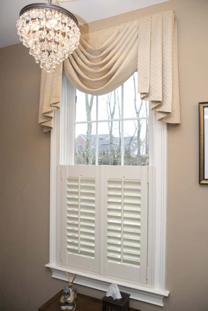 A window with white shutters and a cream-colored, draped valance curtain. Above the window hangs a crystal chandelier, and outside, leafless trees are visible. A small table with a tissue box and decor is below the window.