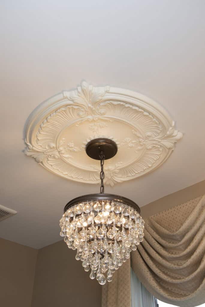 A crystal chandelier with multiple hanging glass droplets is suspended from a decorative white ceiling medallion in a room with beige walls and draped curtains.