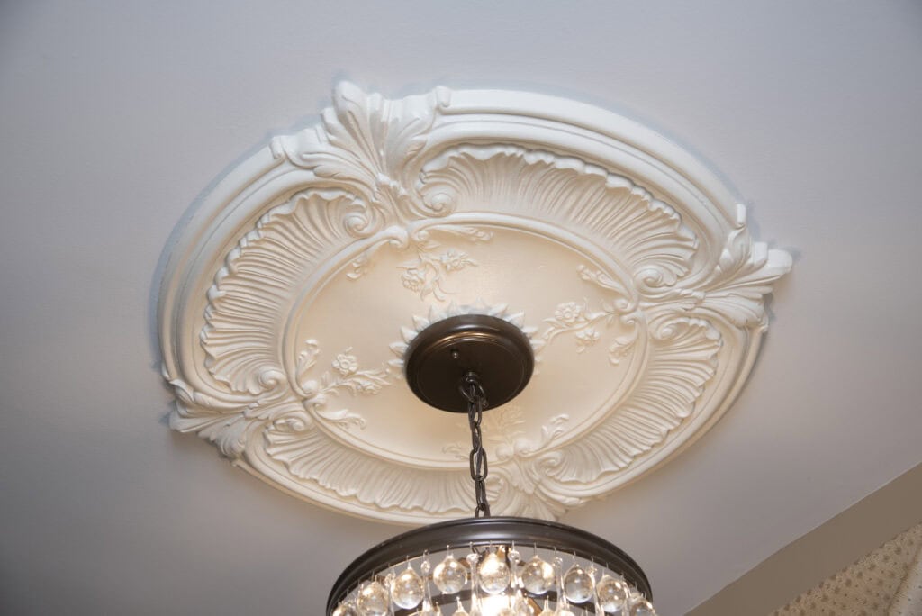 Ornate white plaster ceiling medallion with floral and leaf designs, surrounding the base of a hanging chandelier with crystal accents.