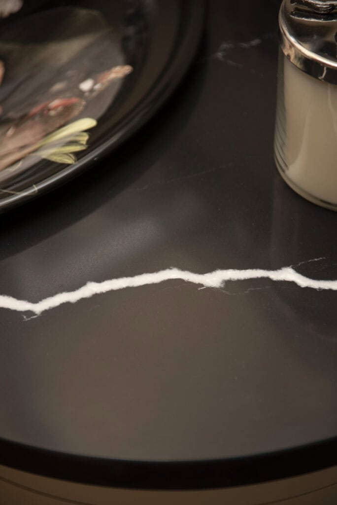 A dark marble surface with a white, jagged vein running through it; a decorative plate and a cylindrical glass jar are partially visible at the edges.
