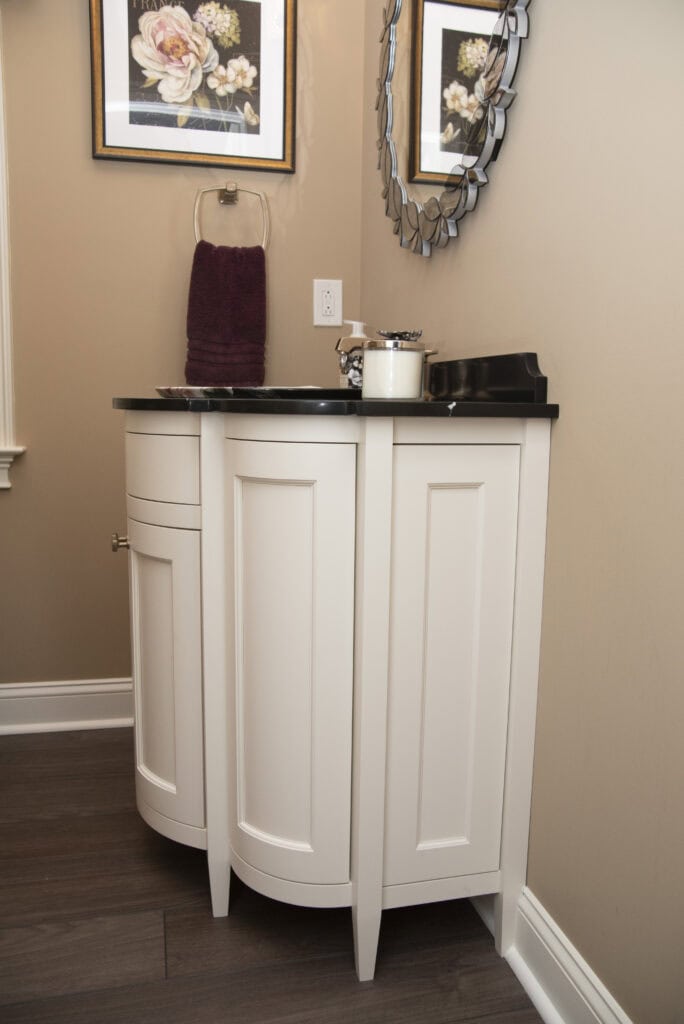 A white corner bathroom vanity with a black countertop, a mounted mirror, a framed floral picture, a purple hand towel, and a cup with toothbrushes on the counter against beige walls and dark wood flooring.