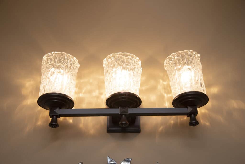 Three wall-mounted light fixtures with textured glass shades emit warm light, casting soft, patterned shadows on a beige wall. The fixture has a dark metal frame and a modern, elegant design.