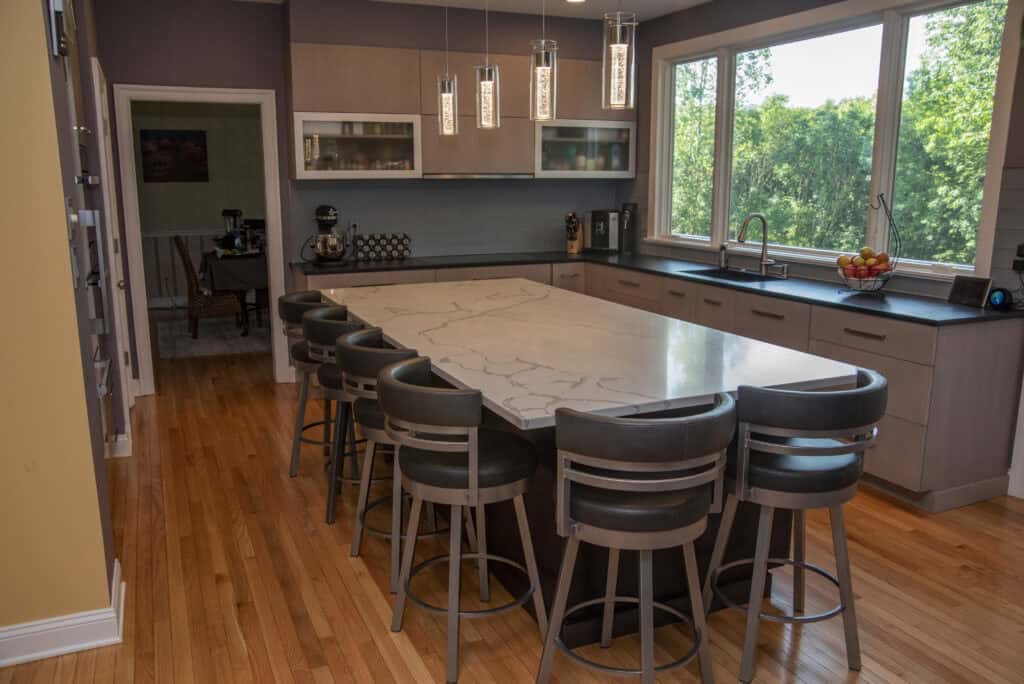 Modern kitchen with a large marble island, six gray chairs, wooden floors, pendant lights, gray cabinets, stainless appliances, a sink under big windows, and a bowl of fruit on the counter. Dining area visible through doorway.