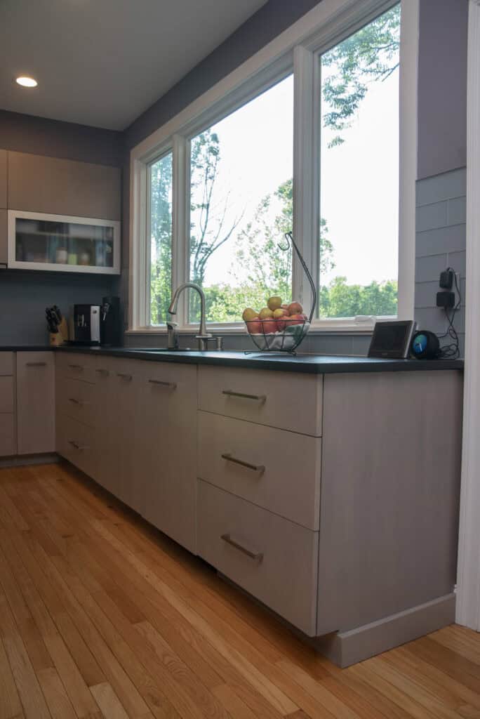 Modern kitchen with light wood cabinets, a large window overlooking greenery, a countertop with a fruit basket and small appliances, and light hardwood floors.