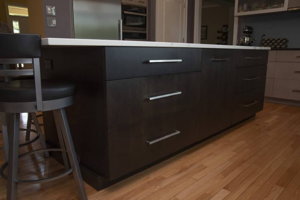 A modern kitchen island with dark wooden drawers, silver handles, and a white countertop sits on a light wood floor next to a black and gray cushioned bar stool.