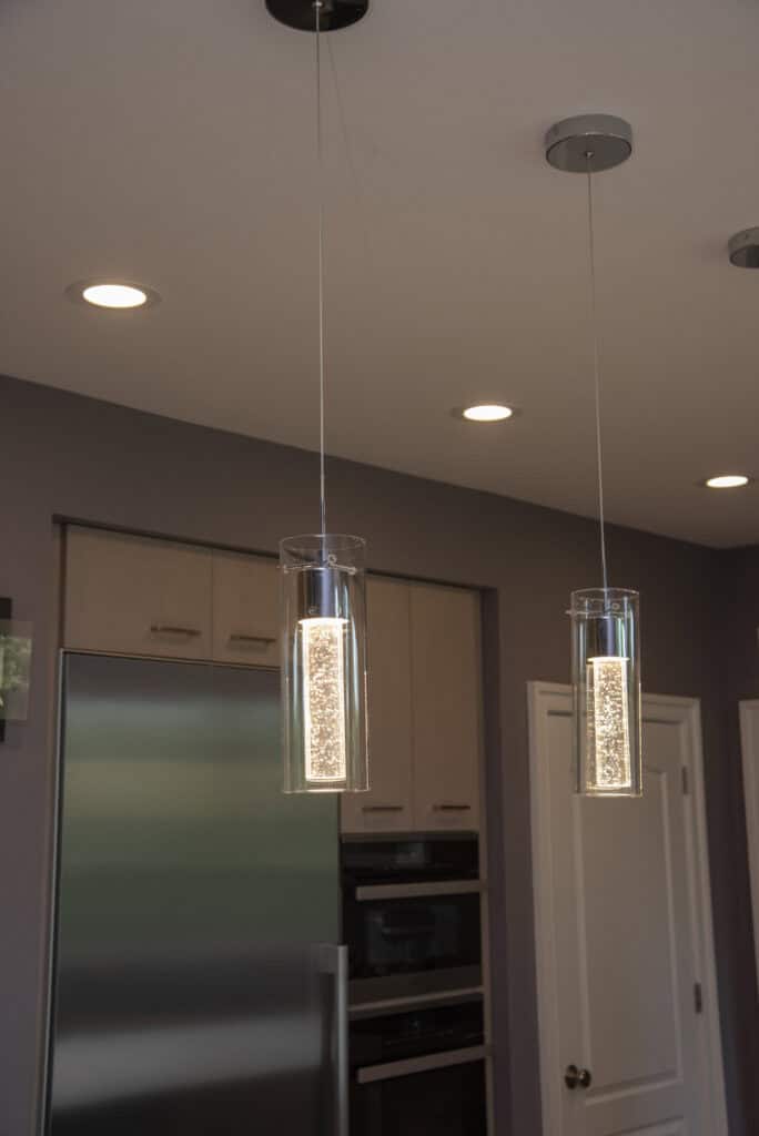 Two modern pendant lights with cylindrical glass covers hang from the ceiling in a kitchen with recessed lighting, stainless steel appliances, and light wood cabinets.
