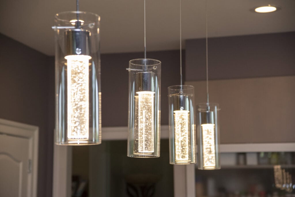 Four modern cylindrical pendant lights with clear glass shades hang from the ceiling, illuminated and glowing warmly in a contemporary kitchen setting. A door and cabinets are visible in the background.