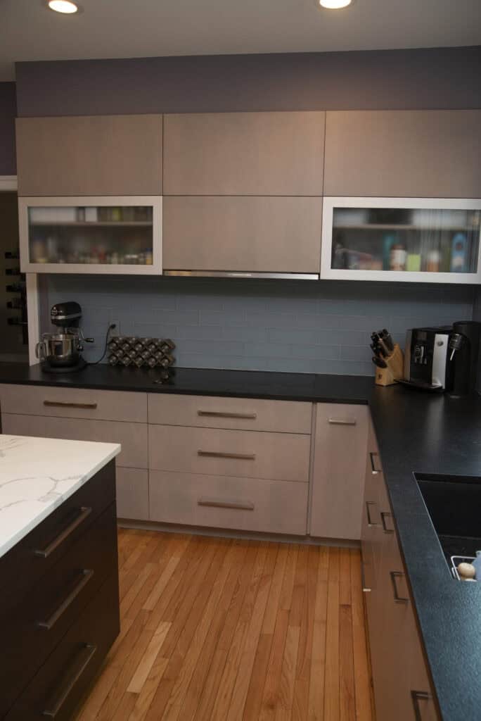 Modern kitchen with light wood cabinets, black countertops, stainless steel appliances, a knife block, wine rack, and a coffee maker on the counter, all set against a light blue tiled backsplash and wood flooring.