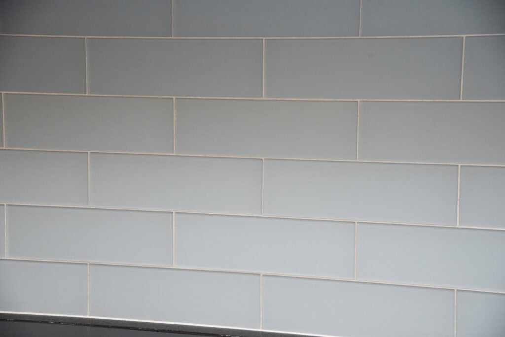 A wall with light gray rectangular tiles arranged in a horizontal subway pattern, separated by thin white grout lines.