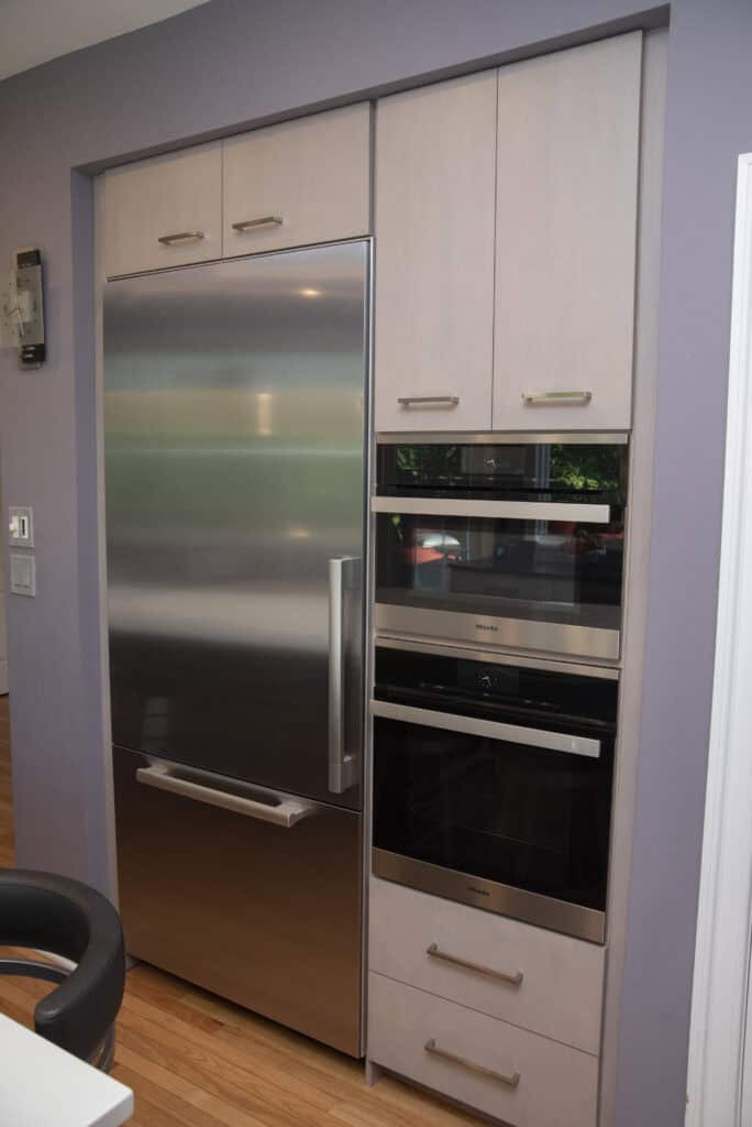 A modern kitchen with stainless steel built-in refrigerator, two stacked wall ovens, light wood cabinetry, and light purple walls. Hardwood flooring and a glimpse of a black chair are also visible.
