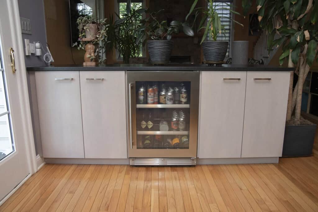 A modern kitchen with light wood cabinets, a glass-door mini fridge filled with cans and bottles, black countertop, potted plants, and hardwood floor.
