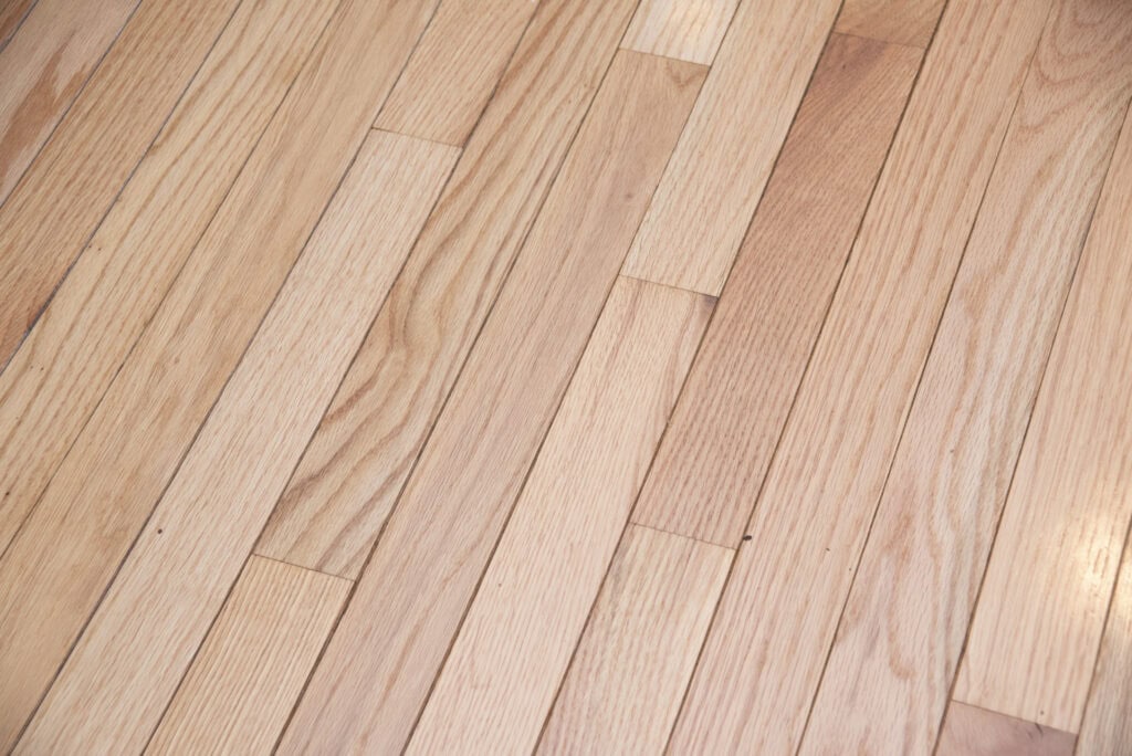 Light-colored wooden floor with visible natural grain and pattern, consisting of narrow, rectangular planks laid side by side in a staggered arrangement. The surface appears clean and well-maintained.
