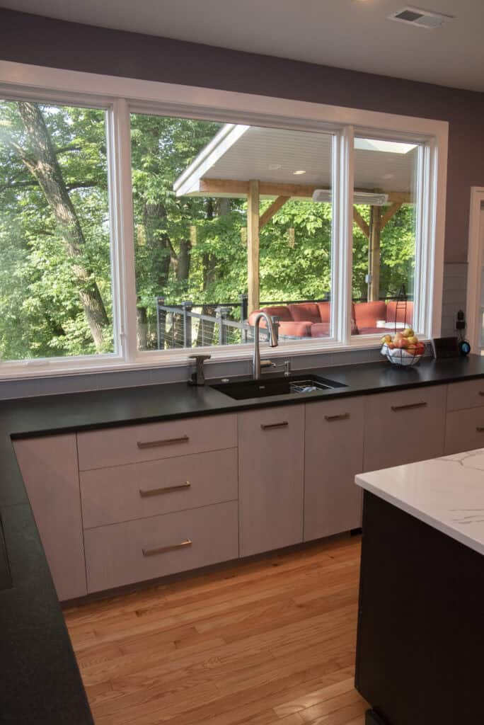 Modern kitchen with light wood cabinets, black countertops, and a large window overlooking a porch with outdoor seating and lush green trees in the background. Natural light fills the room.