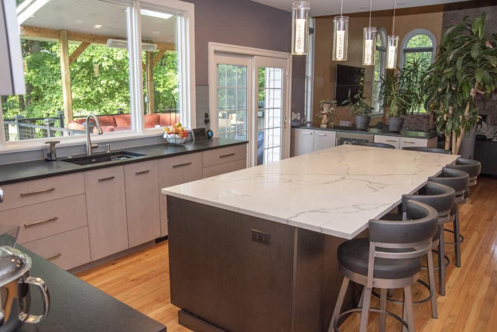 Modern kitchen with a large marble island, black chairs, light wood cabinets, pendant lights, and a view of a green outdoor patio through large windows and a glass door. Several potted plants and fruit bowls decorate the space.