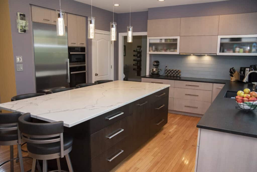 Modern kitchen with a large marble island, four chairs, stainless steel appliances, light wood cabinets, pendant lights, a bowl of fruit, and light wood flooring. The space is tidy and well-lit.