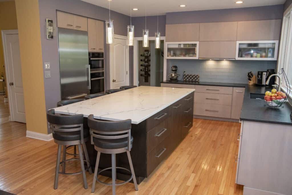 Modern kitchen with a large marble island, three dark chairs, stainless steel appliances, light wood cabinets, pendant lights, and light wood flooring. Fruit bowls and kitchen appliances are visible on the counters.