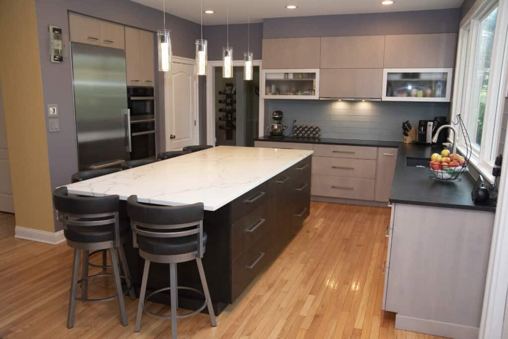 Modern kitchen with a large marble island, four chairs, stainless steel appliances, light wood cabinets, pendant lights, and a bowl of fruit on the counter next to a large window.