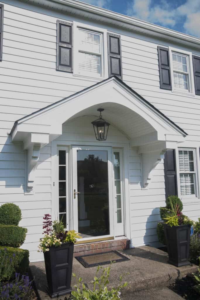 White house with black shutters and a covered front entryway. Glass front door with brick steps, two black planters with greenery and flowers on either side of the entrance, and a lantern-style light above the door.