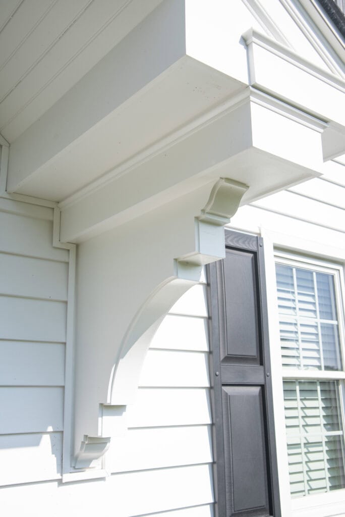Close-up of a white exterior architectural bracket supporting an overhanging roof, attached to a house with white siding and black window shutters. Sunlight casts a shadow of the bracket on the wall.