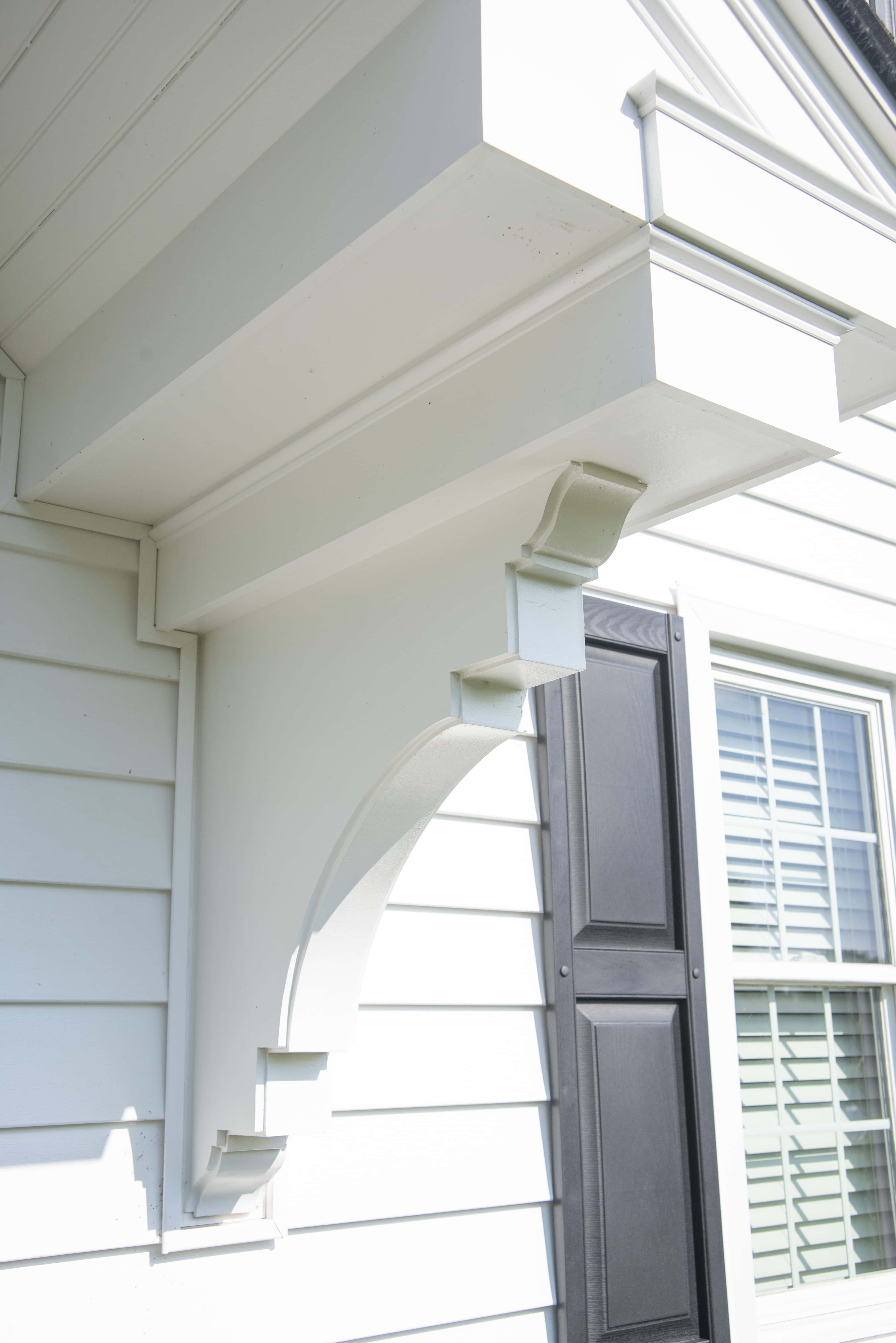 Close-up of a white house exterior shows decorative architectural trim and corbels supporting an overhang, next to black window shutters and white horizontal siding in bright sunlight.