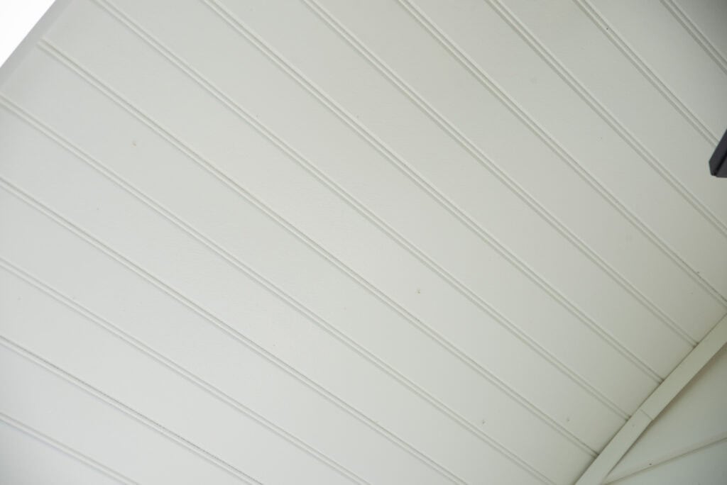 Close-up view of a white, beadboard-paneled ceiling with evenly spaced vertical grooves and a corner edge visible at the bottom right.
