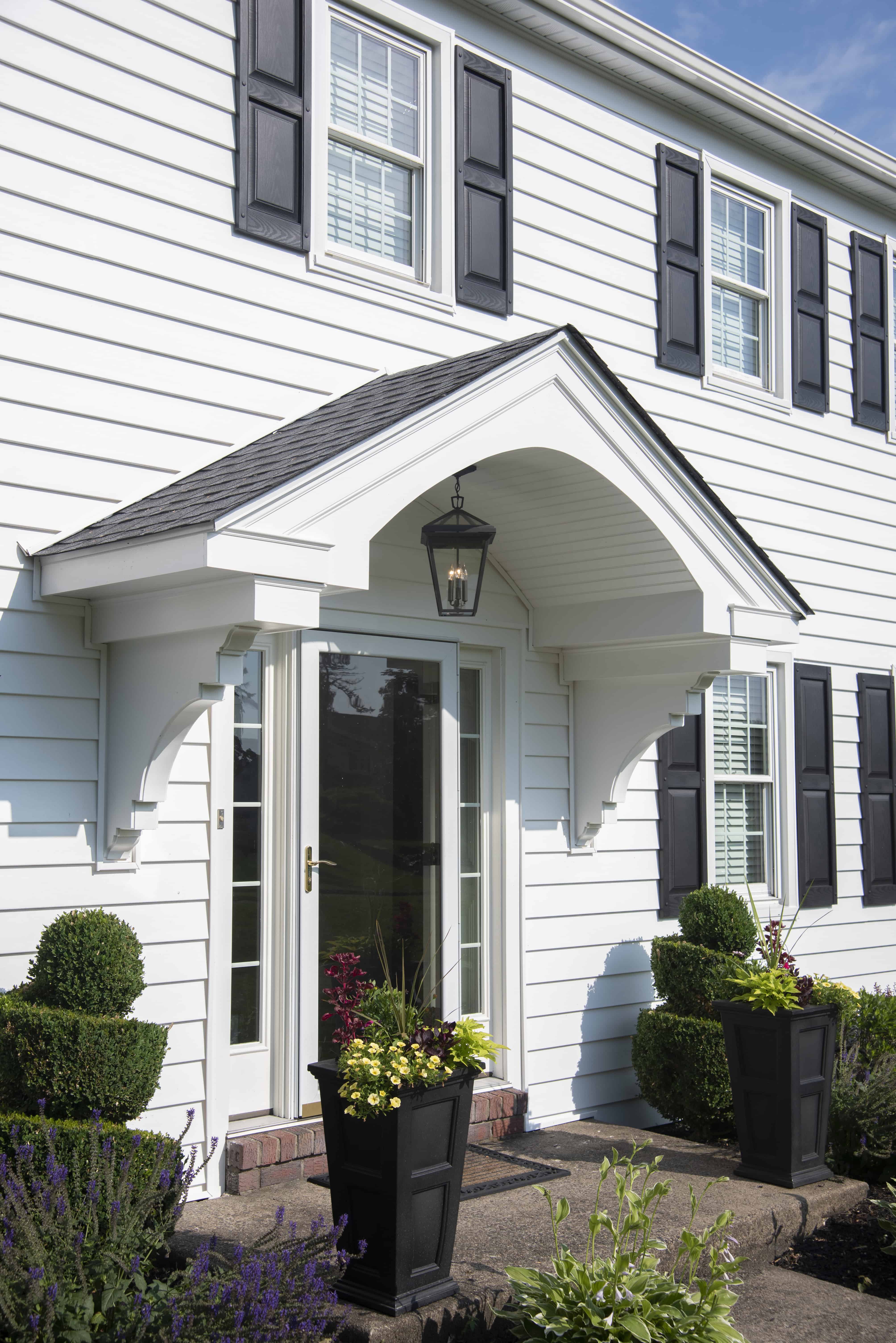 White two-story house with black shutters, a covered front entryway with a glass door, decorative trim, two large potted plants, manicured shrubs, and a lantern hanging above the entrance.
