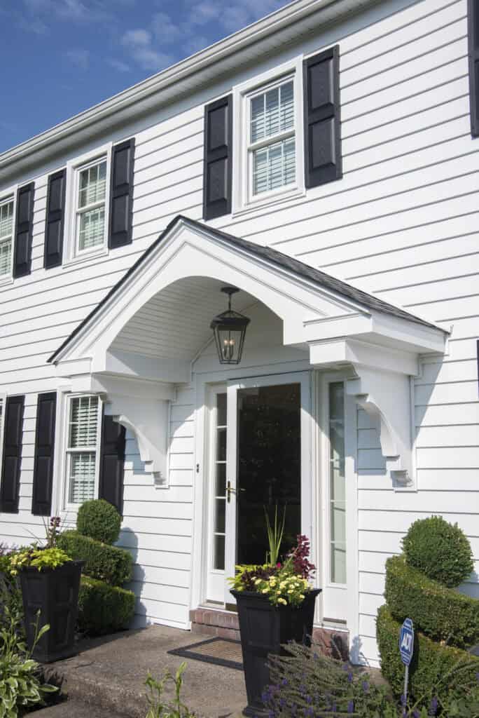 A white two-story house with black shutters, large windows, and a covered entryway. Two black planters with green and purple plants flank the front door. A lantern hangs above the entrance.