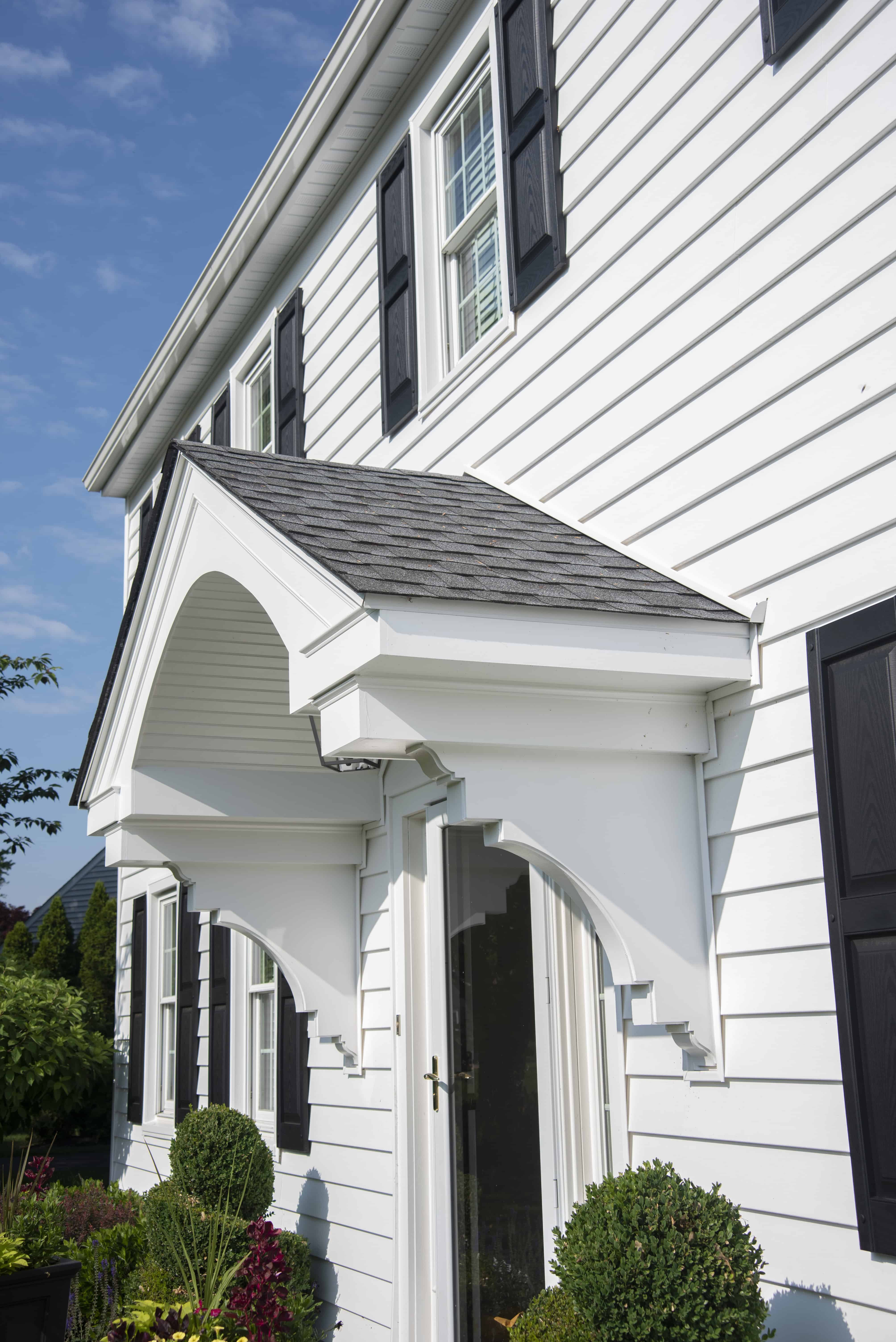 A close-up view of a white house with black shutters, featuring a small covered entryway with decorative trim and neatly trimmed bushes in front. The sky is clear and blue.