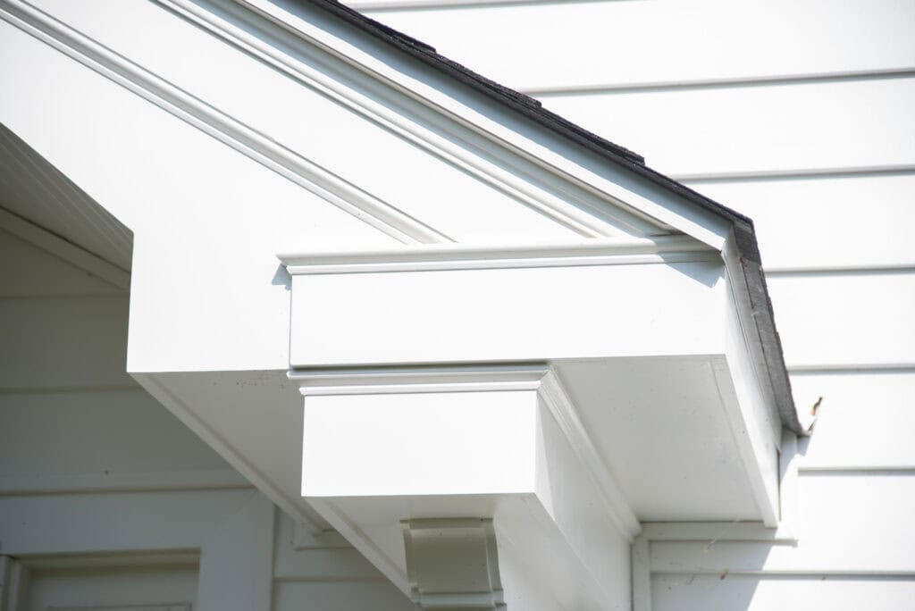 Close-up of the exterior corner of a white house, showing detailed trim and siding beneath a sloped roof with black shingles.