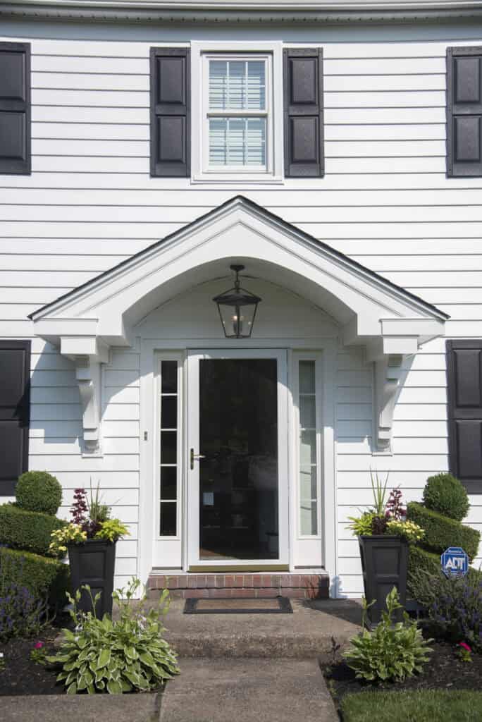 A white house with black shutters and a glass front door. Two large black planters with green and purple plants flank the entrance. A sidewalk leads to the door, and a lantern hangs above the doorway.