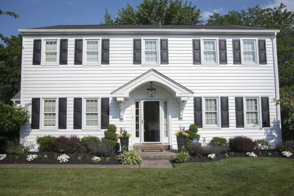 A two-story white house with black shutters, a central front door under a small porch, and a well-maintained garden with flowers and shrubs in front, set against a backdrop of green trees and a blue sky.