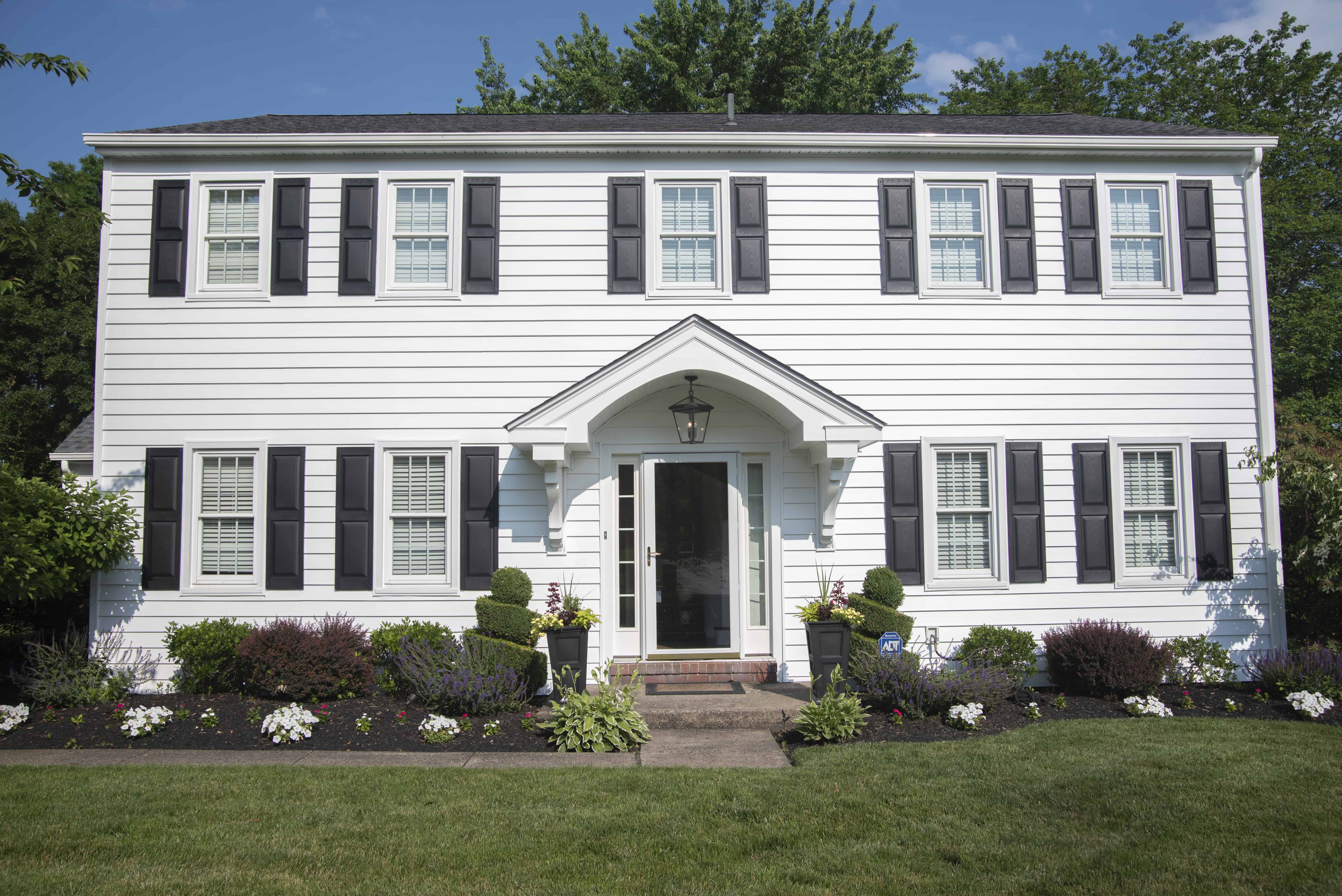 A two-story white house with black shutters, a central front door under a small portico, neatly trimmed bushes, flower beds, and a well-maintained green lawn on a sunny day.