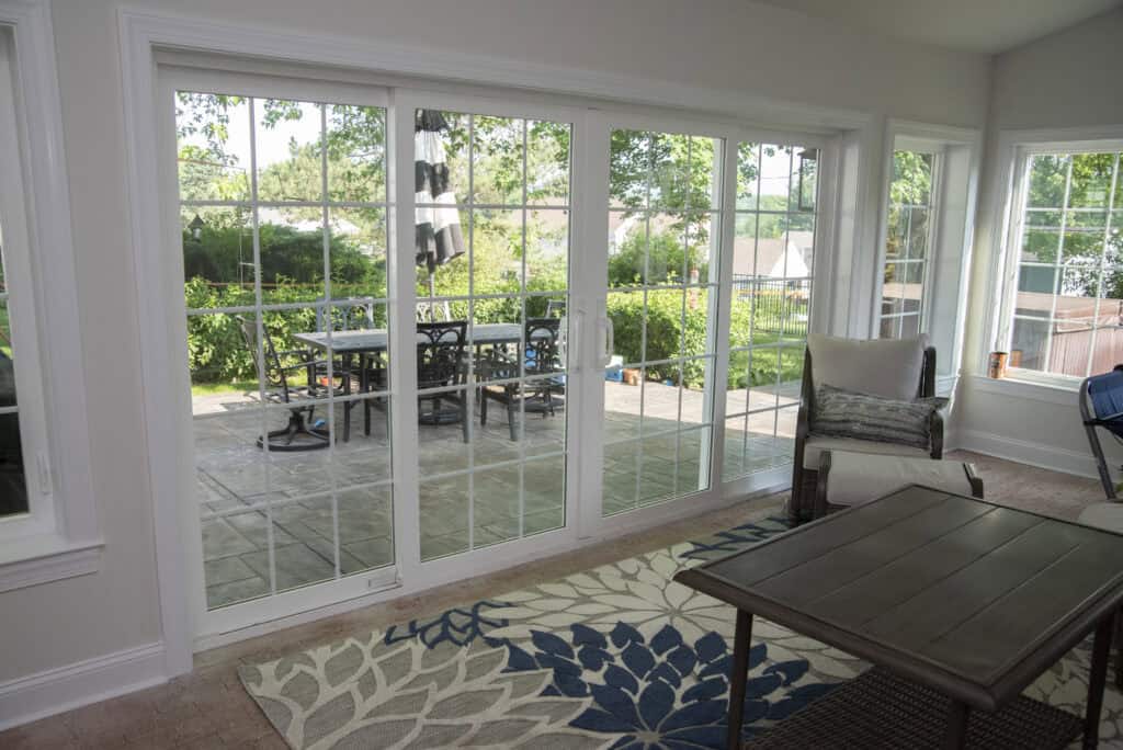A sunroom with large sliding glass doors opens to a patio with a table and chairs. The room has a patterned rug, a coffee table, and a cushioned chair, with green trees visible outside.