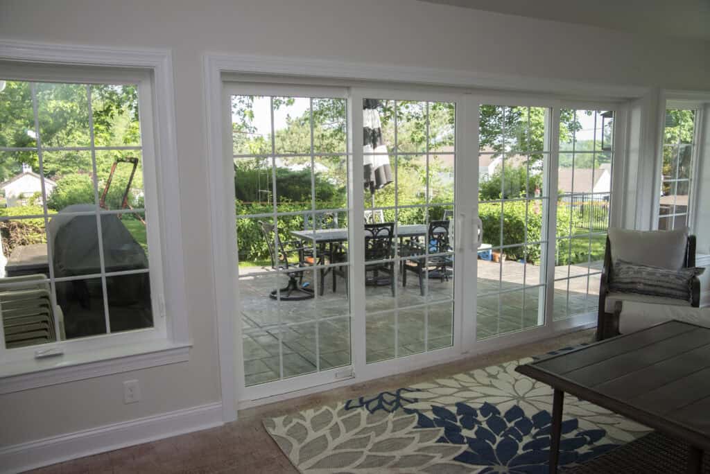 A living room with a patterned rug and a coffee table opens to a patio through large glass sliding doors, revealing an outdoor dining area and lush greenery under a sunny sky.