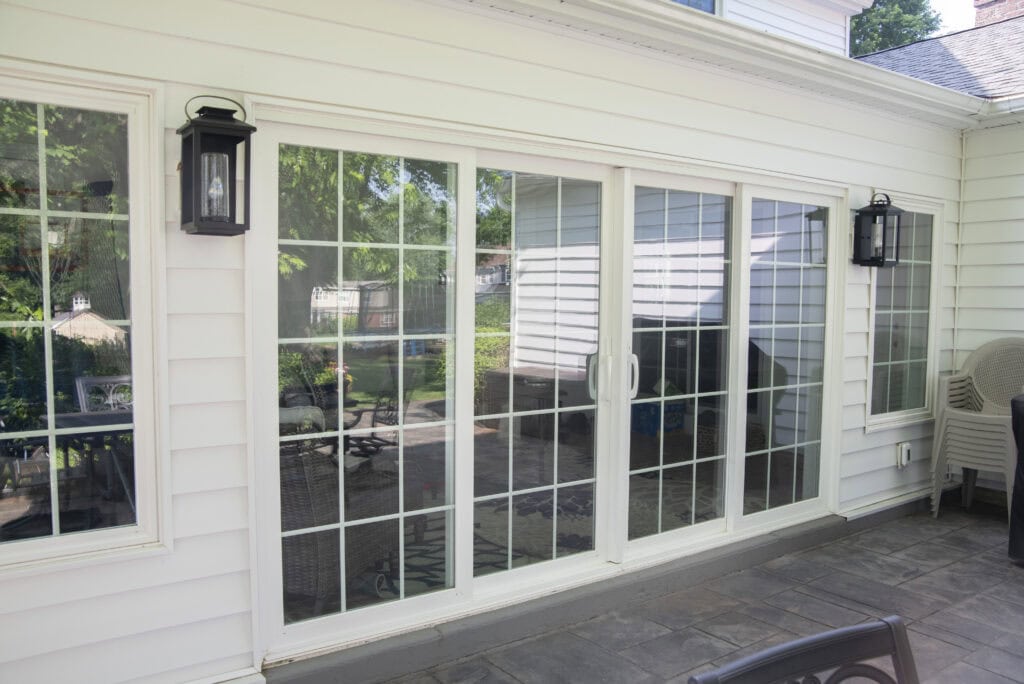 Large white-framed sliding glass doors with grid patterns lead from a patio into a house. Two black outdoor wall lanterns are mounted on either side, and patio furniture is partially visible. The scene is bright and sunny.