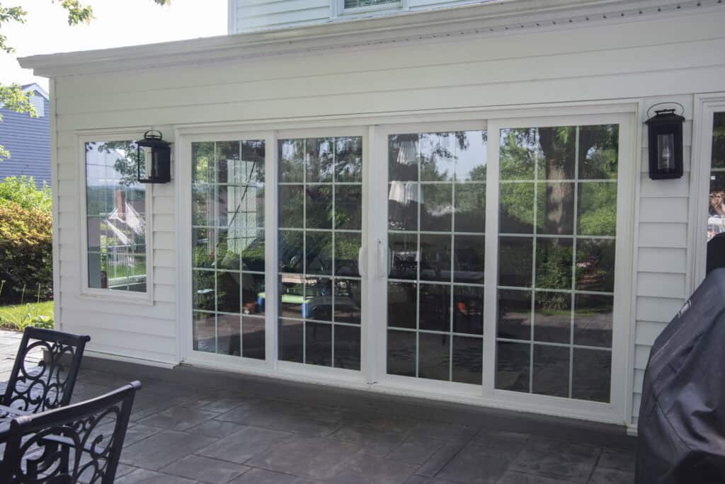 A set of large white sliding glass doors with grid patterns, leading from a patio with stamped concrete flooring into a house. Outdoor furniture and greenery are reflected in the glass.