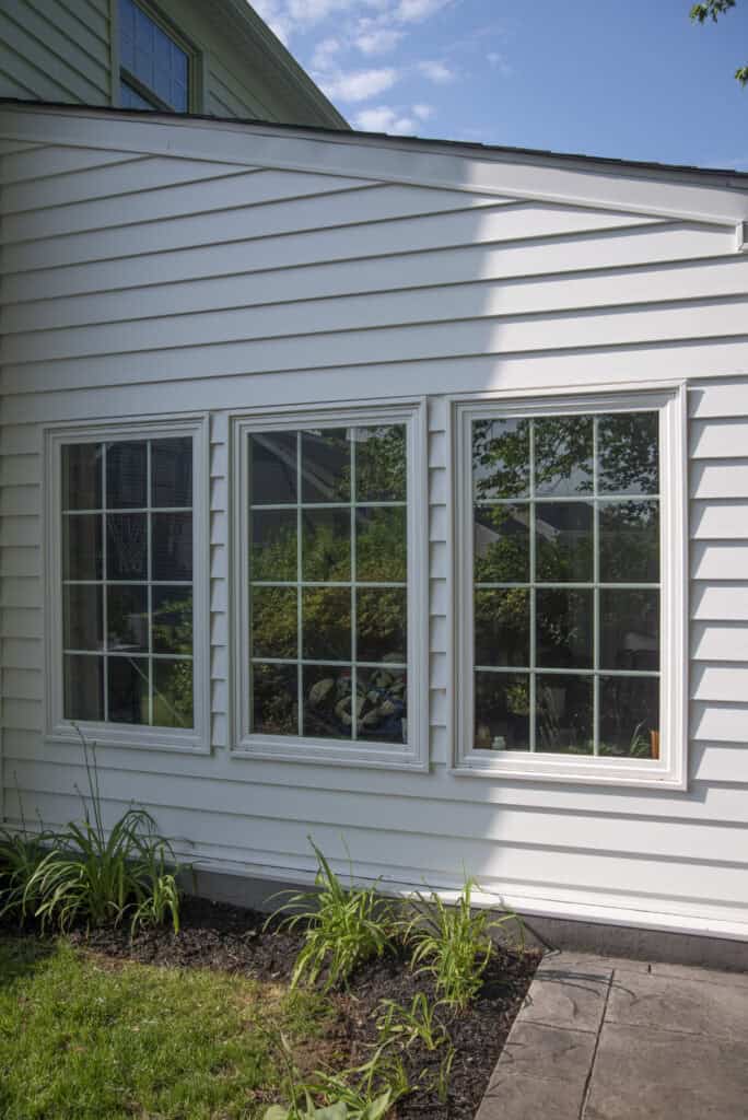 Three large white-framed windows on the side of a house with white siding, reflecting greenery and trees outside. Plants are growing in a garden bed below the windows.