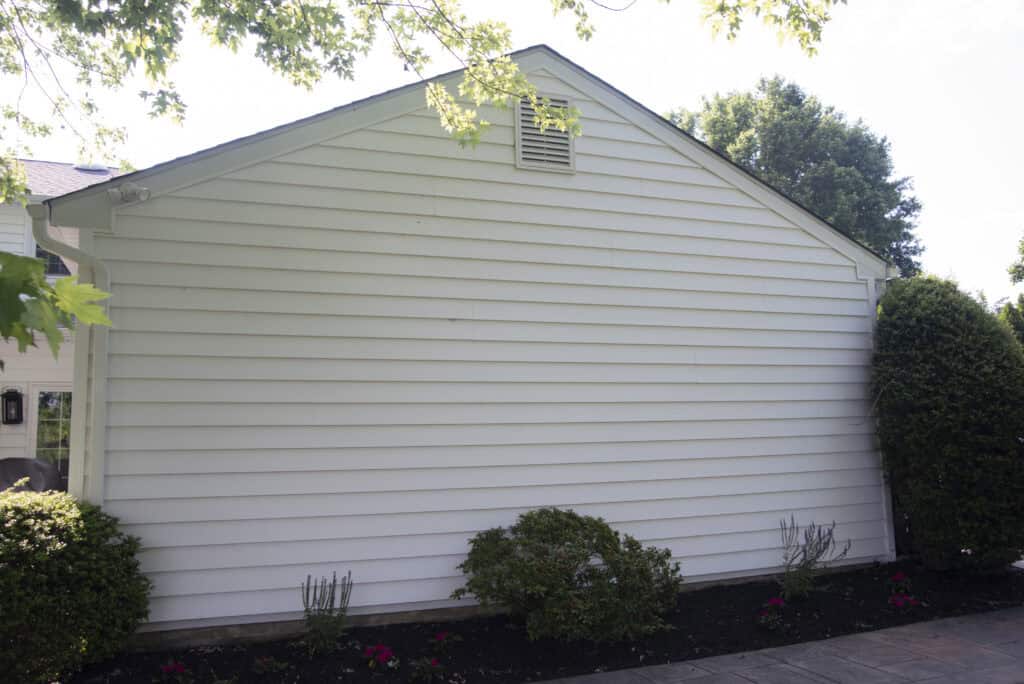 The image shows the exterior side of a white house with horizontal siding. There are a few green bushes, small flowering plants, and a paved walkway in front of the wall. Trees frame the scene from above.