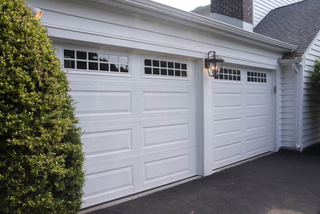 Two white garage doors with decorative windows on a residential home, illuminated by a black outdoor light fixture. A neatly trimmed shrub is on the left, and the driveway is freshly paved.